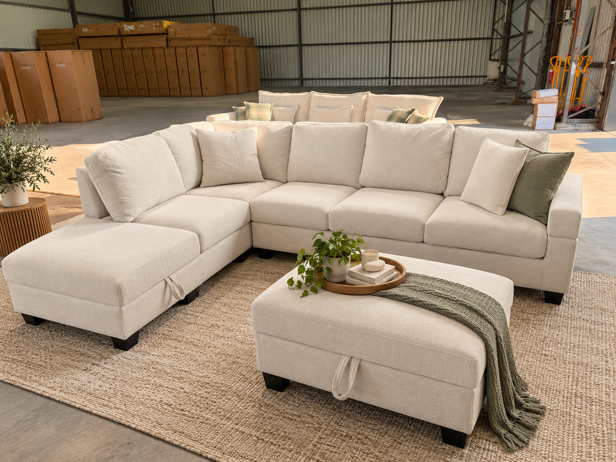 A large sectional sofa in a light beige fabric with matching throw pillows and a matching ottoman, styled in a warehouse showroom setting with a woven rug and a green throw blanket.