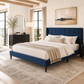 A modern upholstered bed frame in deep blue, styled in a bright, contemporary bedroom with white bedding, beige throw blanket, dark wood nightstands, and a large patterned area rug.