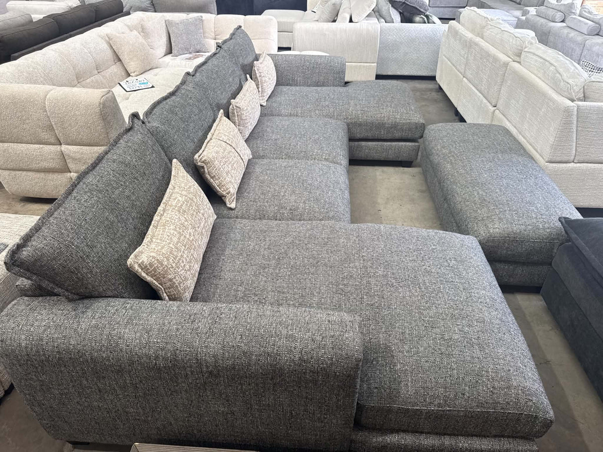 A large gray fabric sectional sofa with a chaise lounge and matching accent pillows, photographed in a showroom with other sofas visible in the background.